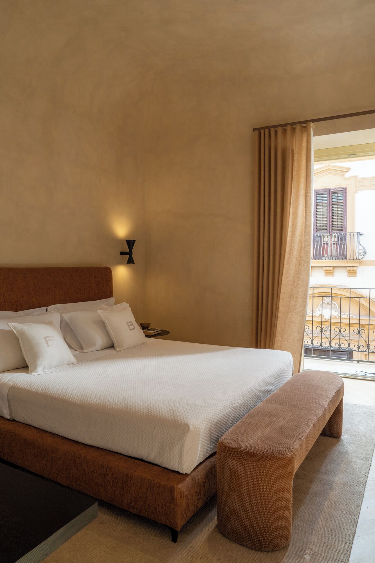 Habitación de hotel con una cama grande, una mesita de noche, un banco y vistas al balcón exterior.