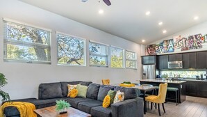 A modern living room with a gray sectional sofa, a wooden coffee table, and a yellow throw blanket. There are large windows with blinds, a kitchen area with dark cabinets, and a colorful wall art piece above the sink. - 5BR Oasis - Pool + Hot Tub + Games - Walk to Fun! (Austin)