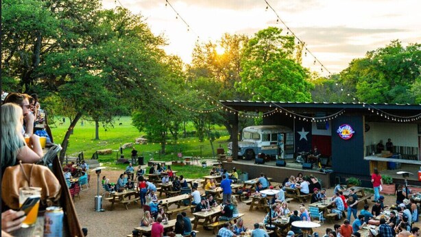 An outdoor dining area with multiple wooden tables and benches, surrounded by trees and a stage with string lights. - 5BR Oasis - Pool + Hot Tub + Games - Walk to Fun! (Austin)