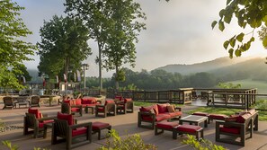 Een terras buiten met houten meubels en rode kussens, omgeven door bomen en uitzicht op een glooiend landschap.