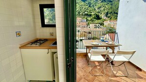 A kitchen with a gas stove and a window overlooking a balcony. The balcony has a table and chairs, and a view of a mountainous landscape.