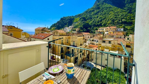 A balcony with a view of a coastal town, featuring buildings with red-tiled roofs and a mountainous backdrop.