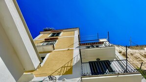 A multi-story building with balconies, a clear blue sky, and a hillside in the background.