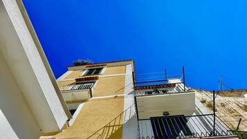 Un edificio de varios pisos con balcones, cielo azul despejado y una colina al fondo.