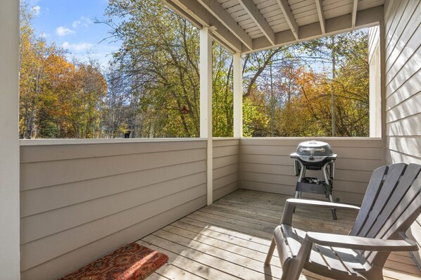 A covered porch with a grill and Adirondack chairs, overlooking a forest with autumn foliage.