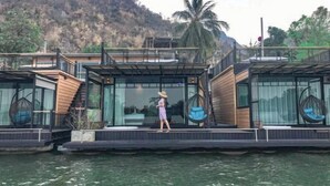 A floating house with a glass facade, wooden deck, and a person walking on the deck. - Blueskyraft Resort (Kanchanaburi)