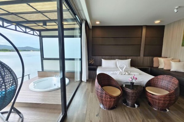 A modern hotel room with a large bed, a sitting area with wicker chairs, a small table, and a view of the water. - Blueskyraft Resort (Kanchanaburi)