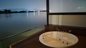 A modern bathroom with a large bathtub, a glass railing, and a view of a calm lake at dusk. - Blueskyraft Resort (Kanchanaburi)
