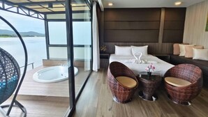A modern bedroom with a large bed, two wicker chairs, a small table, and a view of a body of water through large windows. - Blueskyraft Resort (Kanchanaburi)