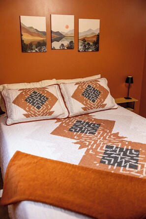 A bed with patterned pillows and a quilt, a bedside table with a lamp, and three landscape paintings on the wall. - Near Wisp & Lake• Private • Fire Pit • Pets & Kids (McHenry)