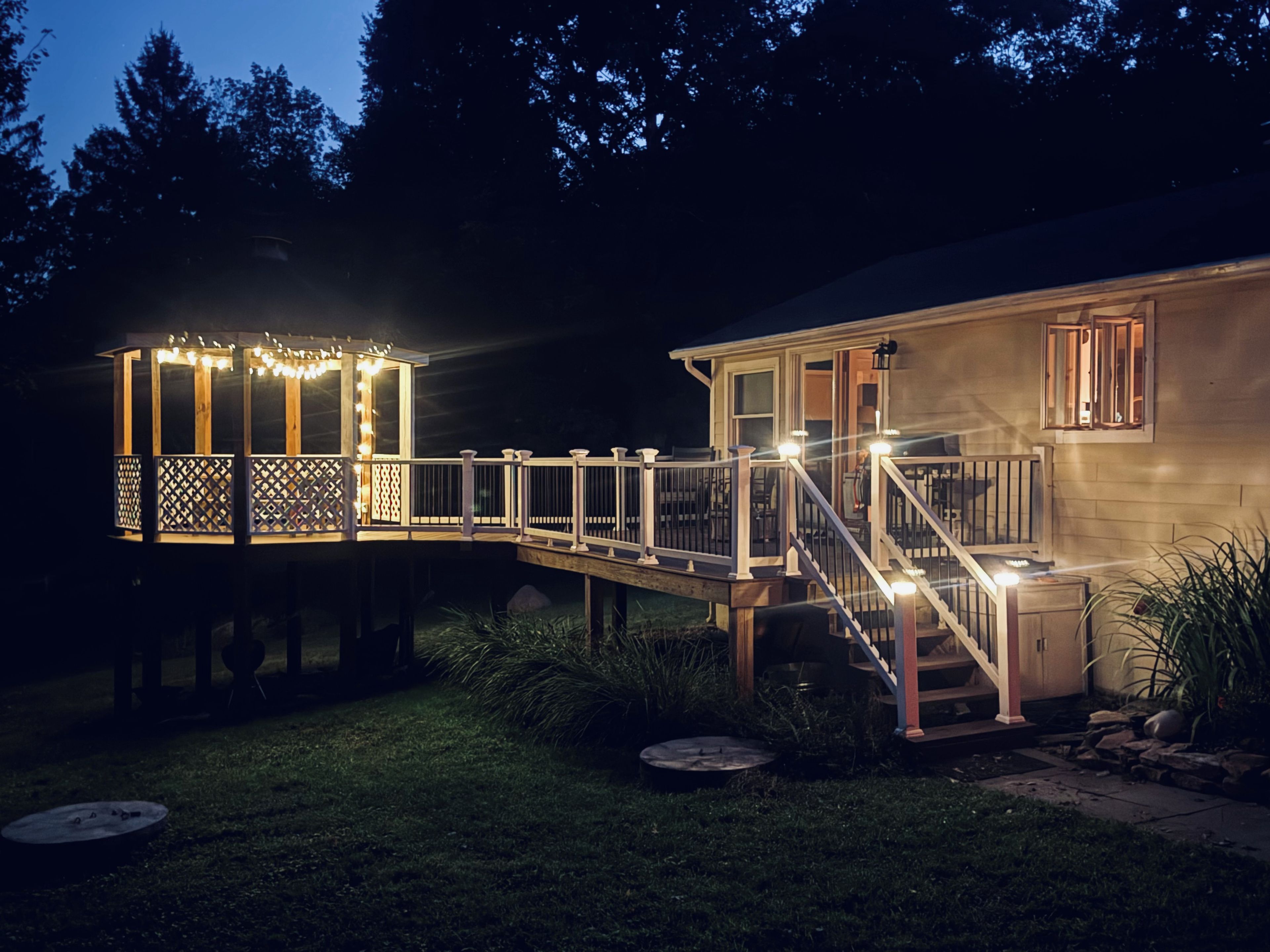 A wooden deck with railings and string lights, leading to a house with a porch and a window.