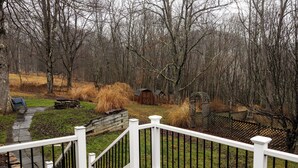 Eine Holzterrasse mit weißer Reling, die auf einen Garten mit Steinmauer, Holzschuppen und einem eingezäunten Bereich mit hohem Gras blickt.