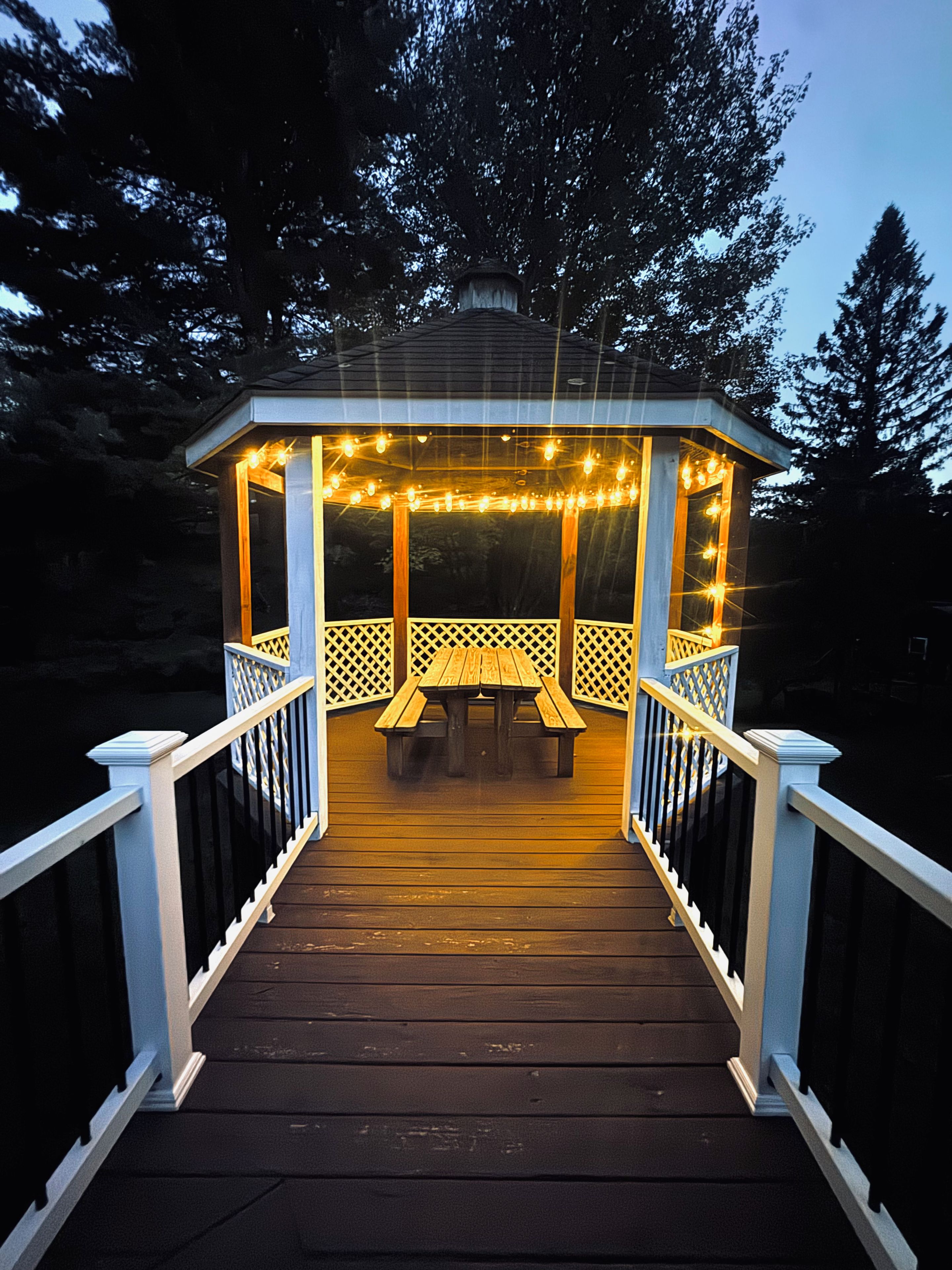 A wooden gazebo with a bench and string lights, surrounded by a deck with railings.