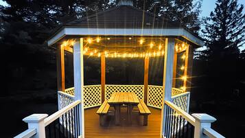 A wooden gazebo with a bench and string lights, surrounded by a deck with railings.