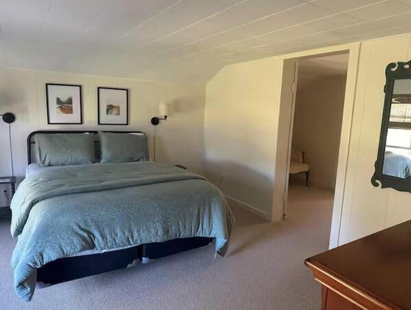 Una camera da letto con un letto, due quadri appesi al muro, un comodino, una cassettiera e una porta che dà su un'altra stanza.