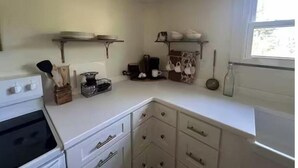 Een kleine keuken met witte kastjes, een fornuis en planken met servies en keukengerei.