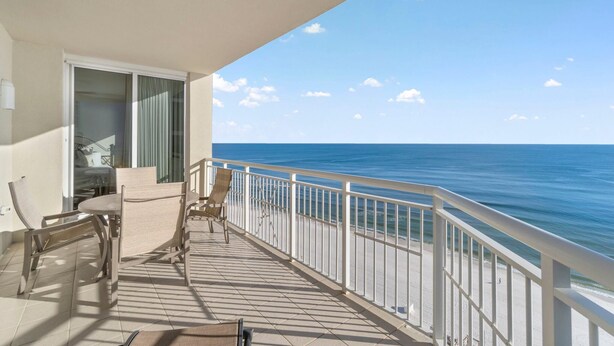 A balcony with a view of the ocean, a table, chairs, and a door leading to a room. - Indigo West 1404 (Pensacola)