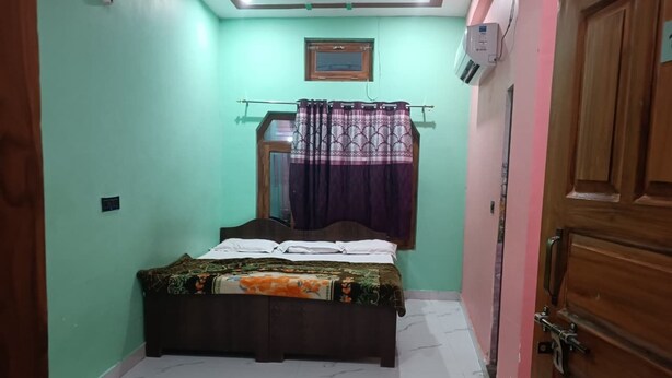 A bedroom with a bed, a window with curtains, a wooden door, and a ceiling fan. - Chandrika Palace - Best Hotel Near Ram Mandir (Ayodhya)