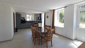 A spacious dining area with a large table and chairs, a kitchen in the background, and large windows providing natural light. - Maison neuve 2 chambres au calme à Ceaulmont (Ceaulmont)