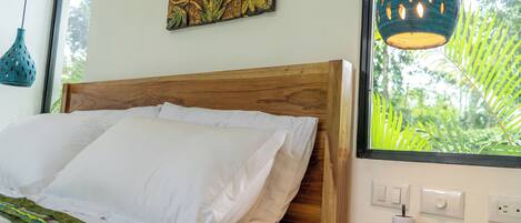 Una cama bien hecha con ropa blanca, un cabecero de madera y una colcha estampada verde. Encima de la cama hay un cuadro colgado en la pared y una lámpara colgante azul.