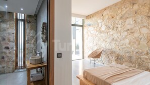 A bathroom with a stone wall, a wooden shelf with a sink, and a window with blinds.