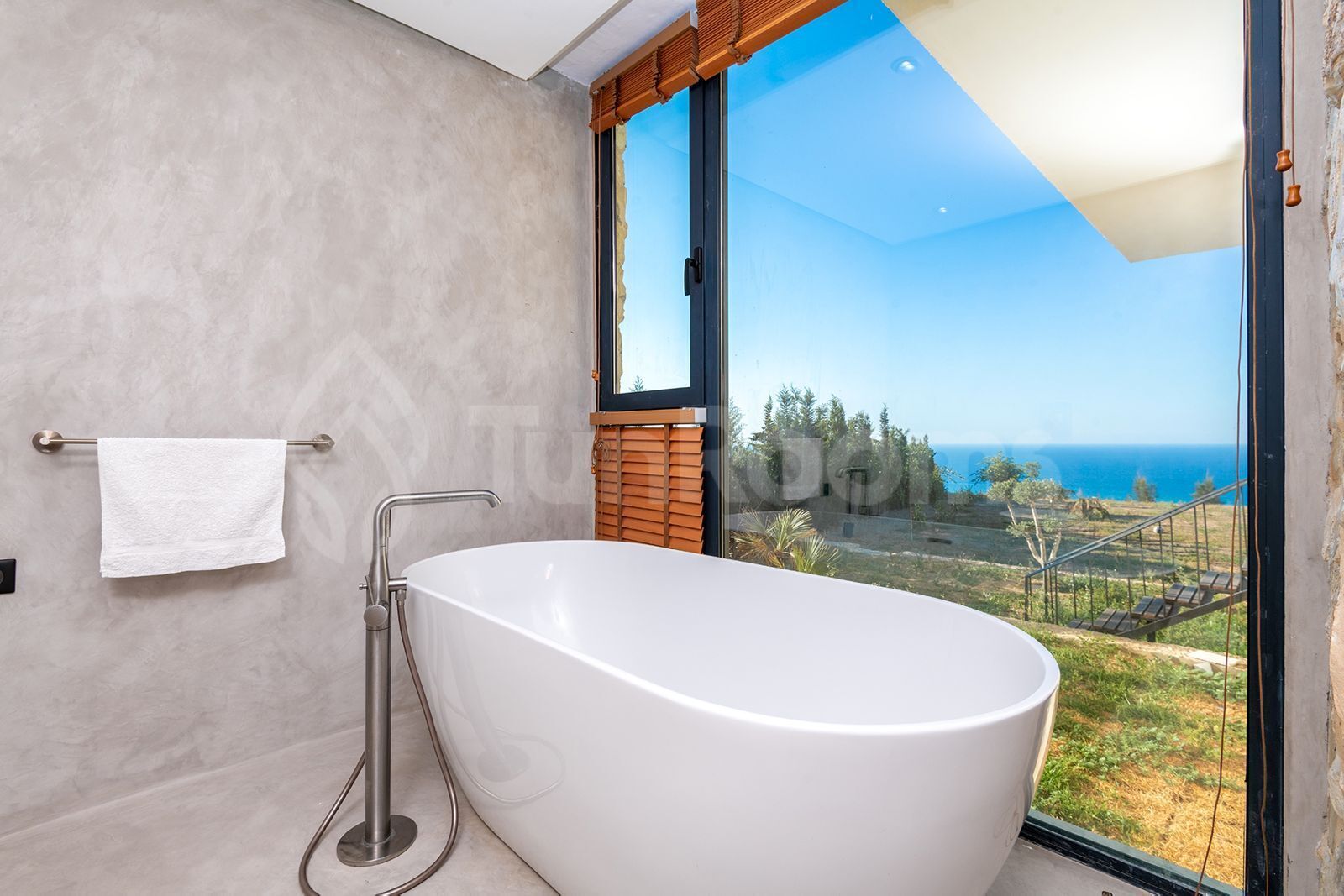 A modern bathroom with a freestanding bathtub, a towel rack, and a large window offering a view of the ocean and greenery.