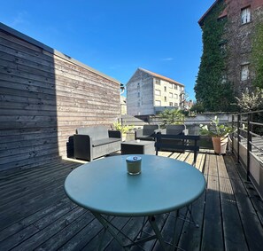 A wooden deck with a round table and chairs, a potted plant, and a building in the background.