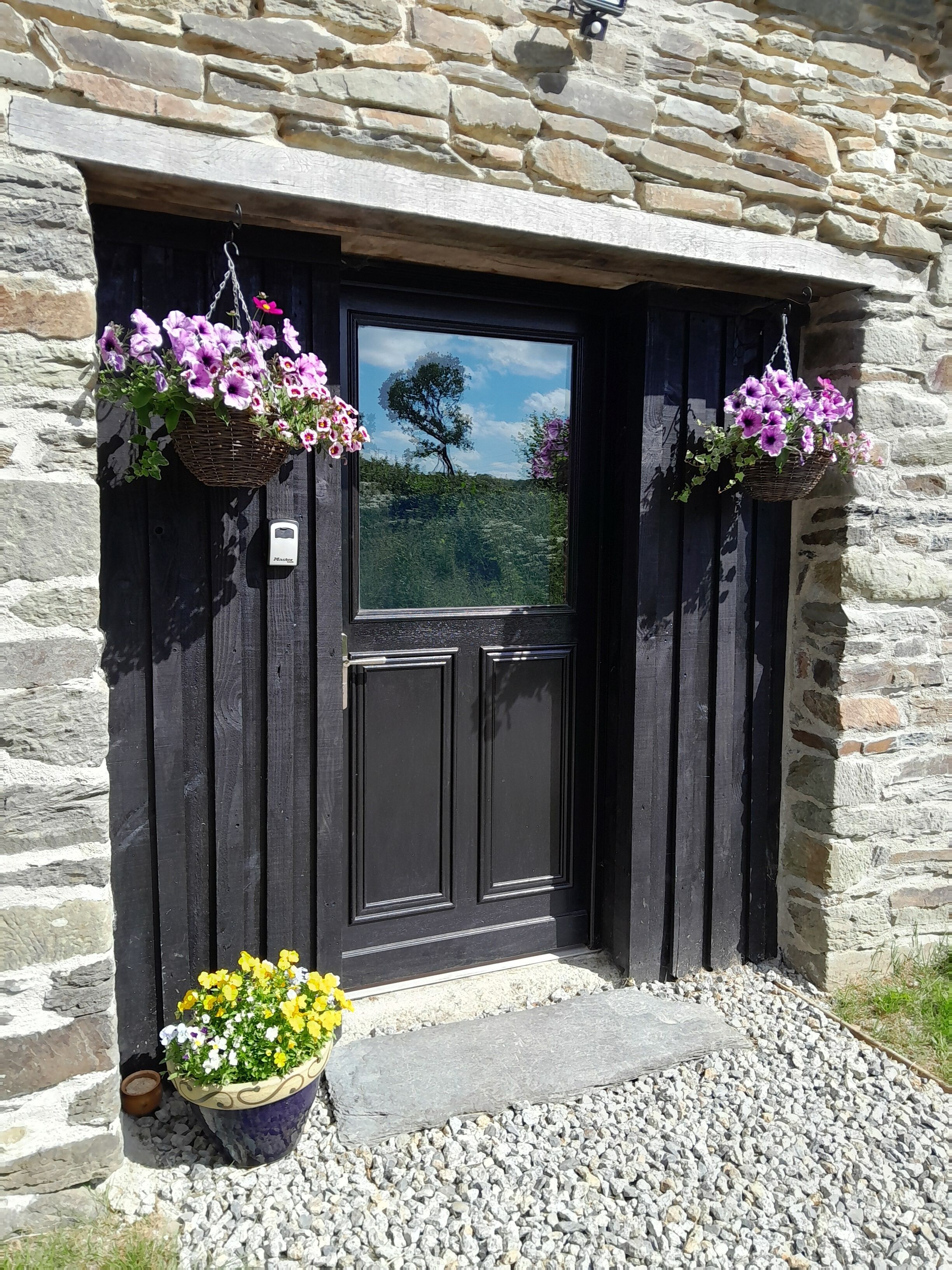 A stone building with a dark wooden door, two hanging flower baskets, and a potted plant with yellow and white flowers.