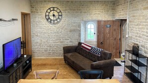 Een woonkamer met een stenen muur, een klok, een houten deur, een raam, een televisie, een bank en een tafel.