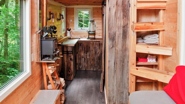 A compact, wooden cabin with a kitchenette, a bed, and a view of the forest.