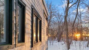 A wooden cabin with large windows, surrounded by snow-covered ground and trees.