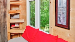 A wooden cabin interior with a bed, shelves, and a window with a view of greenery.
