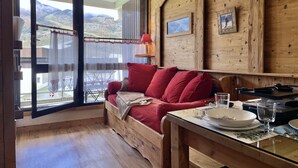 Ein gemütliches Zimmer mit Holzvertäfelung, einer Couch mit roten Kissen, einem Esstisch mit Tellern und Gläsern und Blick auf Berge durch ein großes Fenster.