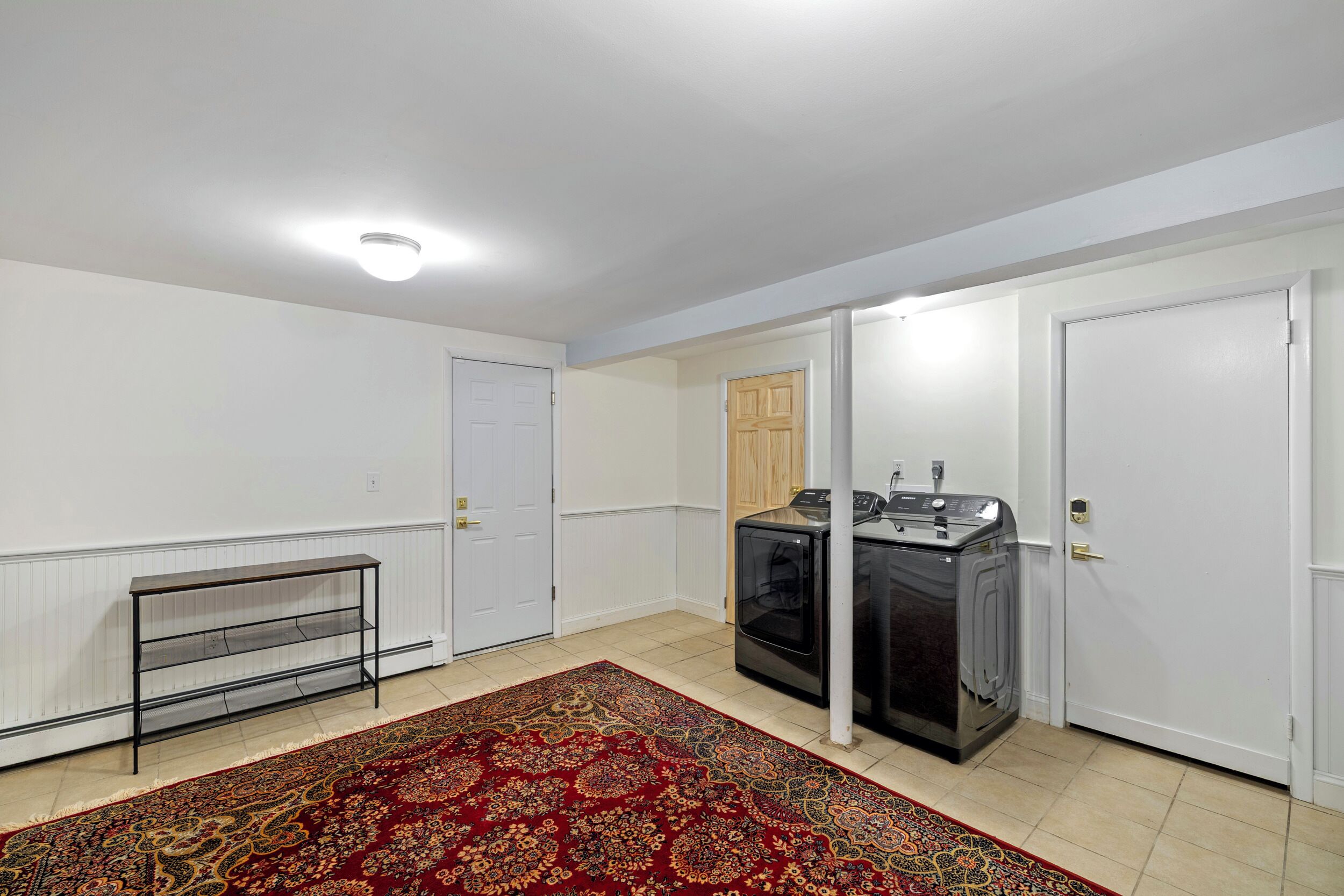 Una habitación con una estantería de metal negro, un tapete rojo estampado, una lavadora y puertas blancas.