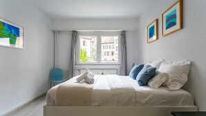 Un lit bien fait, avec du linge de lit blanc, une chaise bleue et une fenêtre avec des rideaux.