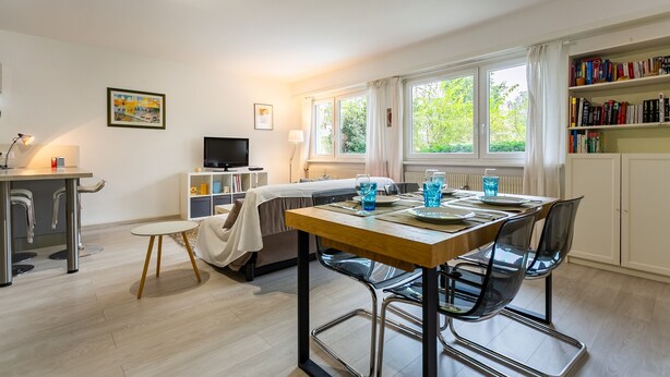 Une pièce moderne et bien éclairée, comprenant une table à manger, des chaises, un canapé, un téléviseur et une bibliothèque.