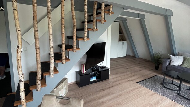 A modern living room with a wooden staircase, a flat-screen TV, and a sofa. - Haus Majda - A place with heart, history and friendship (Ronshausen)
