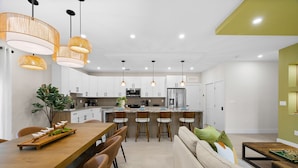 Una cocina moderna con armarios blancos, una isla central, iluminación colgante y una zona de comedor con mesa y sillas de madera.