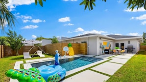 Un patio trasero con piscina, juguetes inflables para la piscina, aro de baloncesto y zona de patio con mobiliario exterior.