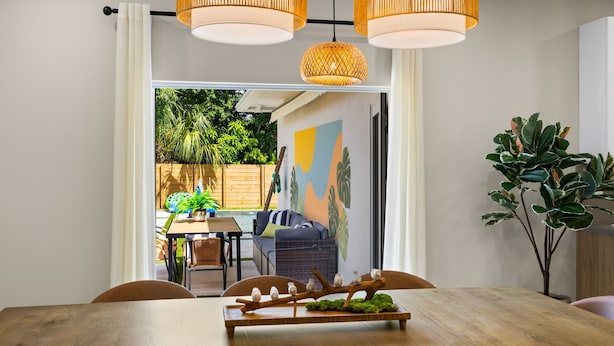 Un comedor con una mesa de madera, sillas y un centro de mesa decorativo con una rama. Se observan sillas de mimbre y un sofá al fondo.