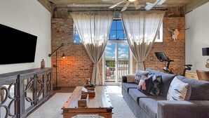 A living room with a brick wall, a large window with curtains, a sofa with cushions, a wooden coffee table, and a flat-screen TV mounted on the wall. - Skyscape - Lofted Luxury Condo Waco/Baylor/Downtown (Waco)