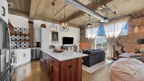 A spacious living area with a large island, a dining table, and a sofa. There is a kitchen with modern appliances and a brick wall. - Skyscape - Lofted Luxury Condo Waco/Baylor/Downtown (Waco)