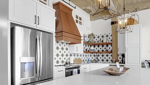 A modern kitchen with a stainless steel refrigerator, wooden cabinets, a large island, and a unique geometric pendant light. - Skyscape - Lofted Luxury Condo Waco/Baylor/Downtown (Waco)