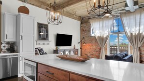 A kitchen with white cabinets, a large island, a flat-screen TV, and a brick wall. - Skyscape - Lofted Luxury Condo Waco/Baylor/Downtown (Waco)