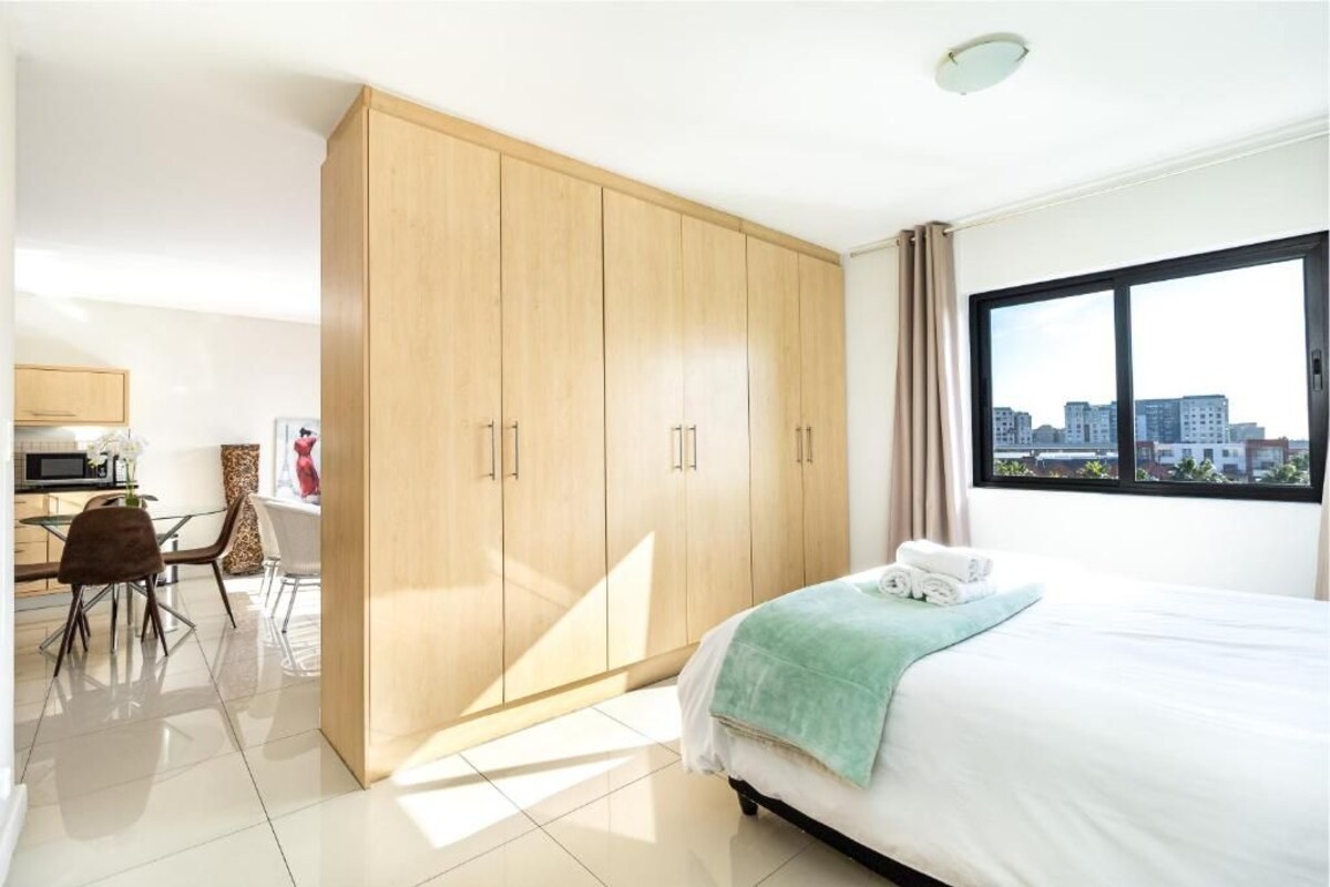 A hotel room with a bed, a large wardrobe, a dining area with chairs, and a view of buildings through a window.