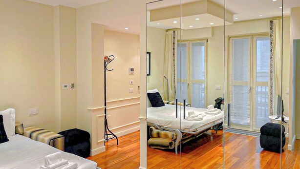 A hotel room with a large mirror, wooden flooring, a bed, bedside tables, and a window with curtains. - Foro Exclusive Suite - 5 mins from Duomo (Milano)