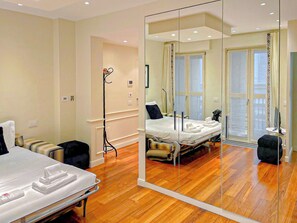 A hotel room with a large mirror, wooden flooring, a bed, bedside tables, and a window with curtains.