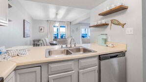 Une cuisine moderne dotée d’un évier à deux bacs, de meubles blancs et d’un lave-vaisselle. Des étagères en bois se trouvent au-dessus de l’évier, où sont disposés des objets décoratifs.