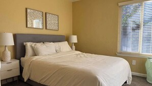 A bedroom with a bed, two bedside tables with lamps, a window with blinds, and two wall art pieces. - Spacious Family Home with Fenced Yard (Modesto)