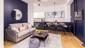 Un salon moderne avec un canapé gris, un miroir rond, un coin repas comprenant une table et des chaises, et une cuisine en arrière-plan.
